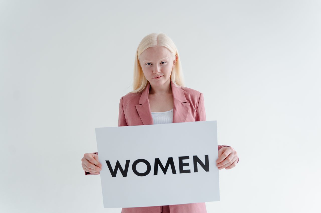 Confident woman in pink blazer holds a sign reading 'Women' on a white background.